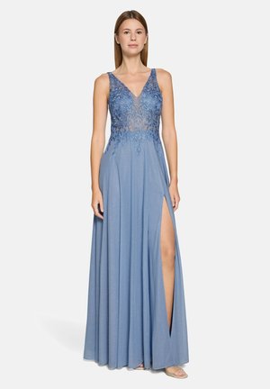 Kobieta stojąca w niebieskiej wieczorowej sukni bez rękawów z koronkowym kwiatowym gorsetem i rozcięciem do uda, o neutralnym wyrazie twarzy, na jednolitym tle.