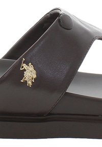 Sandal in pelle marrone con una texture liscia, design open-toe, caratterizzato da un accento logo dorato sulla cinghia. Suola piatta, stile minimalista.