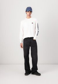 Witte lange mouwen shirt met de tekst "TOMMY JEANS", donkere spijkerbroek en een blauwe pet. Schoenen zijn zwarte sneakers. Model staat tegen een effen achtergrond.