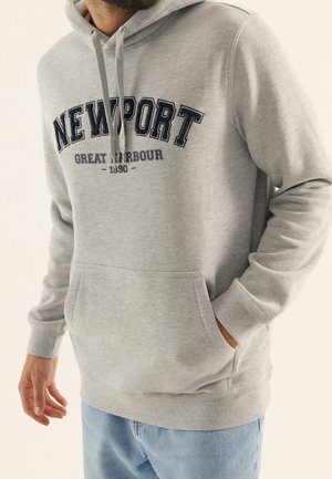 Grijze katoenen hoodie met een vooraanstaande kangoeroezak, een capuchon met trekkoord en marineblauwe letters die "NEWPORT GREAT HARBOUR 1990" zeggen.