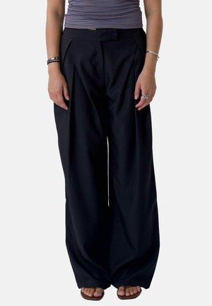 Personne portant un pantalon noir à jambes larges, une chemise rayée, des bracelets, une bague et des sandales ouvertes à l'avant, debout devant un fond uni.