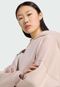 Light pink oversized hoodie with a soft texture, large hood, and visible stitching details. Model's arms are crossed, showcasing the sleeve design.
