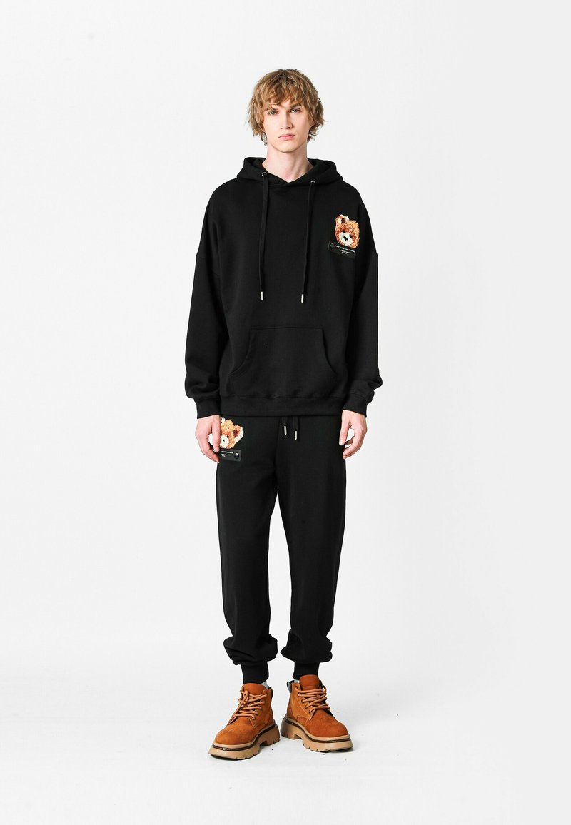 Sudadera con capucha negra y pantalones de chándal de forro polar, con un parche de oso en el pecho y el bolsillo. Botas de trabajo marrones completan el atuendo.