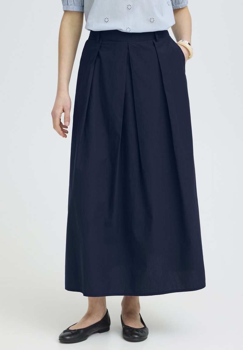 Donna che indossa una gonna blu navy lunga alla caviglia con pieghe, abbinata a ballerine nere e una camicetta azzurra a maniche corte.