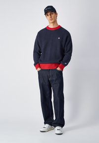 Pull en maille côtelée bleu marine avec un col rouge, associé à un jean sombre et des baskets blanches. Comprend un logo sur la poitrine et des manches contrastantes.
