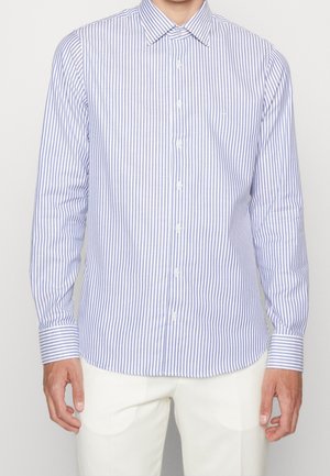 Muž v dlouhém rukávu, košili na knoflíky s vertikálními modrými a bílými pruhy, doplněnou bílými kalhotami.