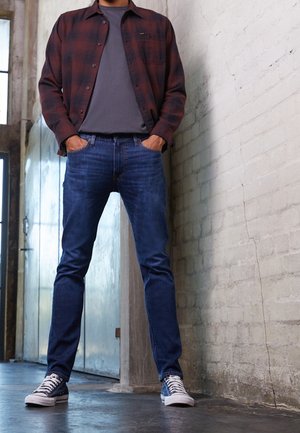 Homme debout à l'intérieur portant un jean bleu, des baskets bleu marine, un t-shirt gris et une chemise à carreaux rouge déboutonnée, les mains dans les poches.