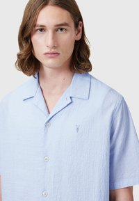Lichtblauw shirt met korte mouwen van een gestructureerde stof, knoopsluiting aan de voorkant en een klein geborduurd logo bij de borst.