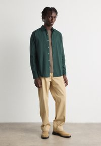 Camisa verde de botones sobre un suéter de punto color beige, combinada con pantalones beige y botas tan. Tela suave con una textura sutil.