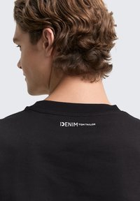Black cotton T-shirt with a round neckline, featuring a white logo that reads “DENIM TOM TAILOR” on the back near the neck.