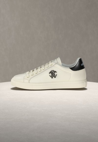 Baskets basses blanches avec languette de talon noire, emblème de logo sur le côté, et lacets blancs, présentées sur une surface et un fond gris neutre.