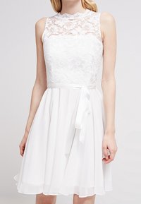 Vit klänning med spetsliv, hög halsringning och en flödande chiffonkjol. Inkluderar ett matchande band som knyts i midjan.