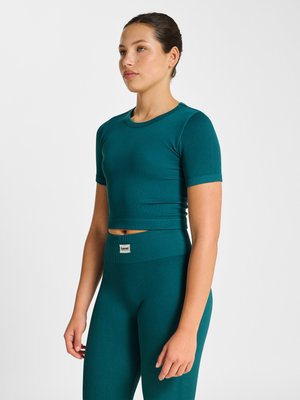 Vrouw die zijwaarts staat, draagt een teal geribbeld crop top met korte mouwen en bijpassende hoge taille leggings tegen een effen achtergrond.