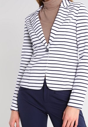 Donna che indossa un blazer bianco con righe orizzontali blu navy, dolcevita beige e pantaloni blu navy su uno sfondo semplice.