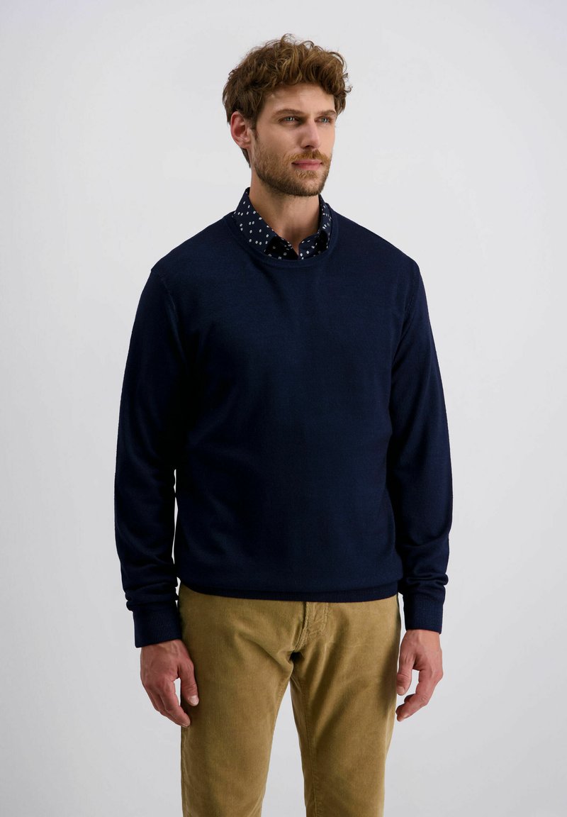 Mann mit lockigem Haar, der einen marineblauen Pullover über einem schwarzen Hemd mit weißen Punkten und beigen Cordhosen trägt, steht vor einem schlichten Hintergrund.