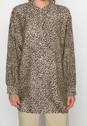 Kvinde iført en løs, langærmet beige bluse med sort uregelmæssigt prikmønster og beige bukser, stående mod en ensfarvet baggrund.