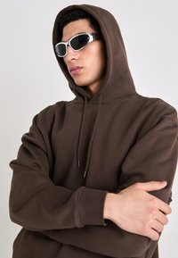 Hnedá mikina s kapucňou na šnúrku a voľným strihom, doplnená o elegantné biele slnečné okuliare. Hladká látka s rebrovanými manžetami a lemom.