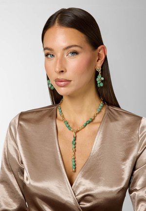 Vrouw met steil bruin haar, die groene kralen oorbellen en bijpassende ketting met gouden accenten draagt, gekleed in een glanzende taupe wikkelblouse.