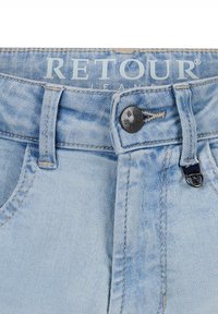 Jeans in denim azzurro con chiusura a bottone, dotati di una vita cucita, passanti per cintura e una piccola etichetta con il logo RETOUR.