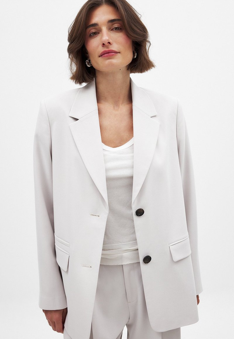 Gai pelēks piegrieztas formas žakete, izgatavota no gludas auduma, ar neizteiksmīgām atlokiem, divu pogu aizdari un sānu kabatām.