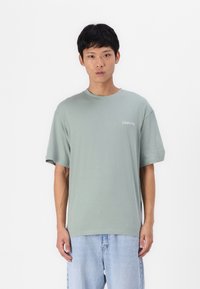 Tricou din bumbac verde deschis, cu o croială lejeră, guler rotund și mâneci scurte. Are un mic logo alb "Club1990" pe piept.
