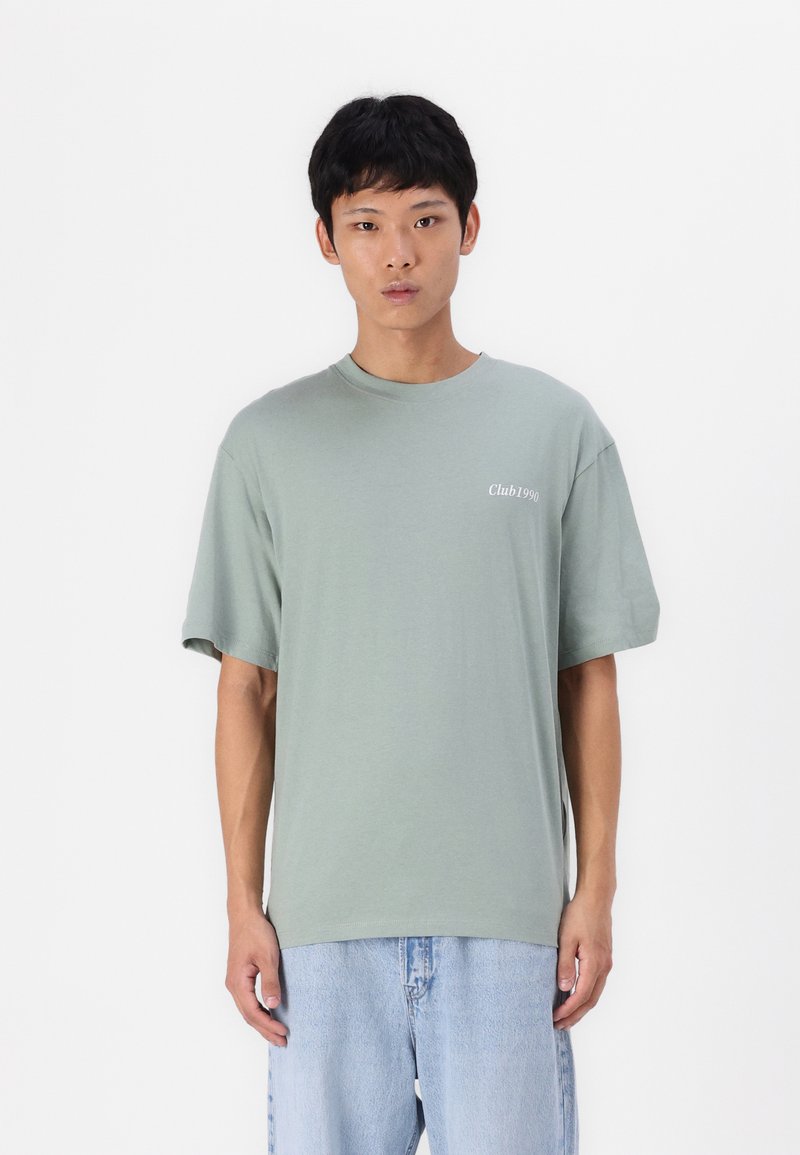 Tricou din bumbac verde deschis, cu o croială lejeră, guler rotund și mâneci scurte. Are un mic logo alb "Club1990" pe piept.