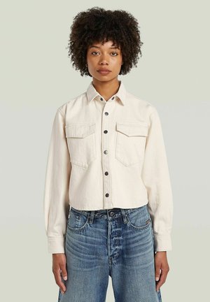 Vrouw met krullend haar, gekleed in een lichtbeige doorgestikte jas met twee borstzakken en een blauwe spijkerbroek, staand tegen een effen achtergrond.