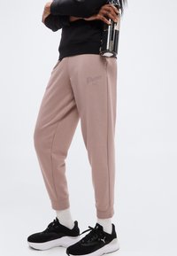 Άτομο που φοράει μωβ φόρμα Puma και μαύρα αθλητικά παπούτσια, κρατώντας ένα μαύρο και διάφανο παγούρι νερού σε λευκό φόντο.