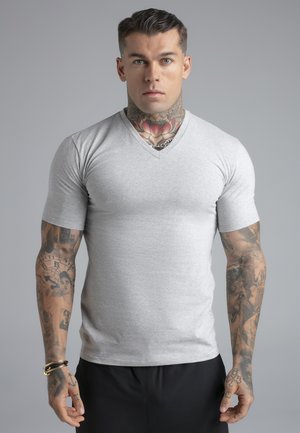 Uomo con tatuaggio floreale sul collo, numerosi tatuaggi sulle braccia e piercing, indossa una maglietta grigia a scollo a V e pantaloni neri, in piedi davanti a uno sfondo grigio uniforme.