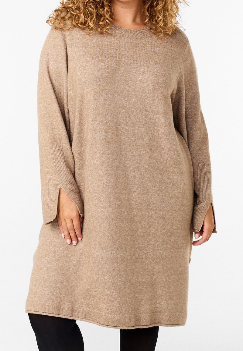 Personne aux cheveux bouclés portant une robe en tricot beige à manches longues et larges avec des trous pour les pouces, debout devant un fond blanc.