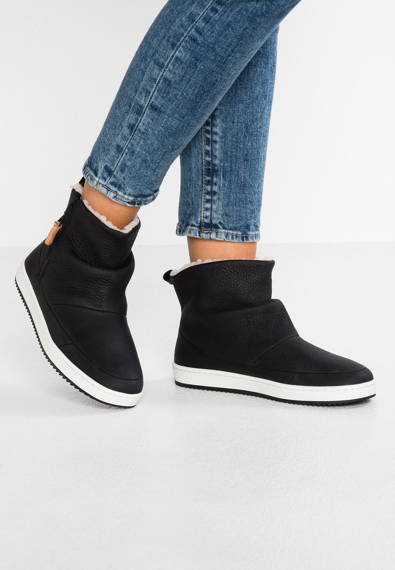 Botas de tobillo de cuero negro con suela de goma blanca, que presentan un acabado texturizado, forro de pelo y un detalle de cremallera en el lateral.