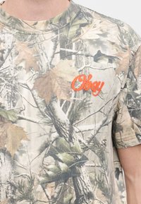 Κοντομάνικη μπλούζα καμουφλάζ με σχέδιο εμπνευσμένο από τη φύση, με φύλλα και δέντρα, κατασκευασμένη από απαλό ύφασμα, με πορτοκαλί κεντημένο λογότυπο "Obey" στο στήθος.