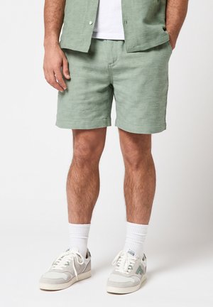 Man draagt lichtgroene gestructureerde korte broek, bijpassend shirt met korte mouwen, witte sokken en witte sneakers met groene accenten.