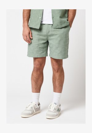 Man draagt lichtgroene gestructureerde korte broek, bijpassend shirt met korte mouwen, witte sokken en witte sneakers met groene accenten.