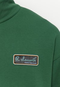 Groene poloshirt van een gestructureerde stof, met een zwart rechthoekig label met witte en lichtblauwe tekst: "R. Lacoste TENNIS HERITAGE."