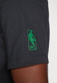 Czarnoszary t-shirt z krótkim rękawem, z zieloną naszywką logo NBA na lewym rękawie. Materiał wydaje się miękki i gładki w dotyku.