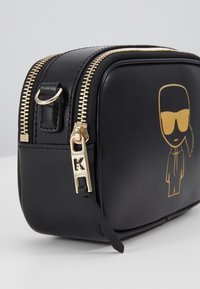 Pochette in pelle nera con texture liscia, zip dorata e grafica dorata di una figura con occhiali da sole. Presenta un dettaglio hardware a forma di 'K'.