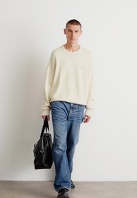 Κρεμ oversized πουλόβερ με διακριτικό λογότυπο, συνδυασμένο με ανοιχτό γαλάζιο τζιν παντελόνι και μαύρη δερμάτινη τσάντα, φορεμένο με μαύρα παπούτσια.