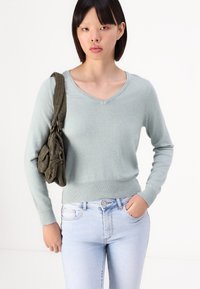 Svijetlozeleni pulover s v-neck izrezom i dugim rukavima, s rebrastim rubom, uparen sa svijetloplavim trapericama. Model drži zelenu torbu preko ramena.