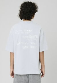 T-shirt surdimensionné gris clair à manches courtes, comportant un texte motivant manuscrit blanc au dos. Texture de tissu douce.