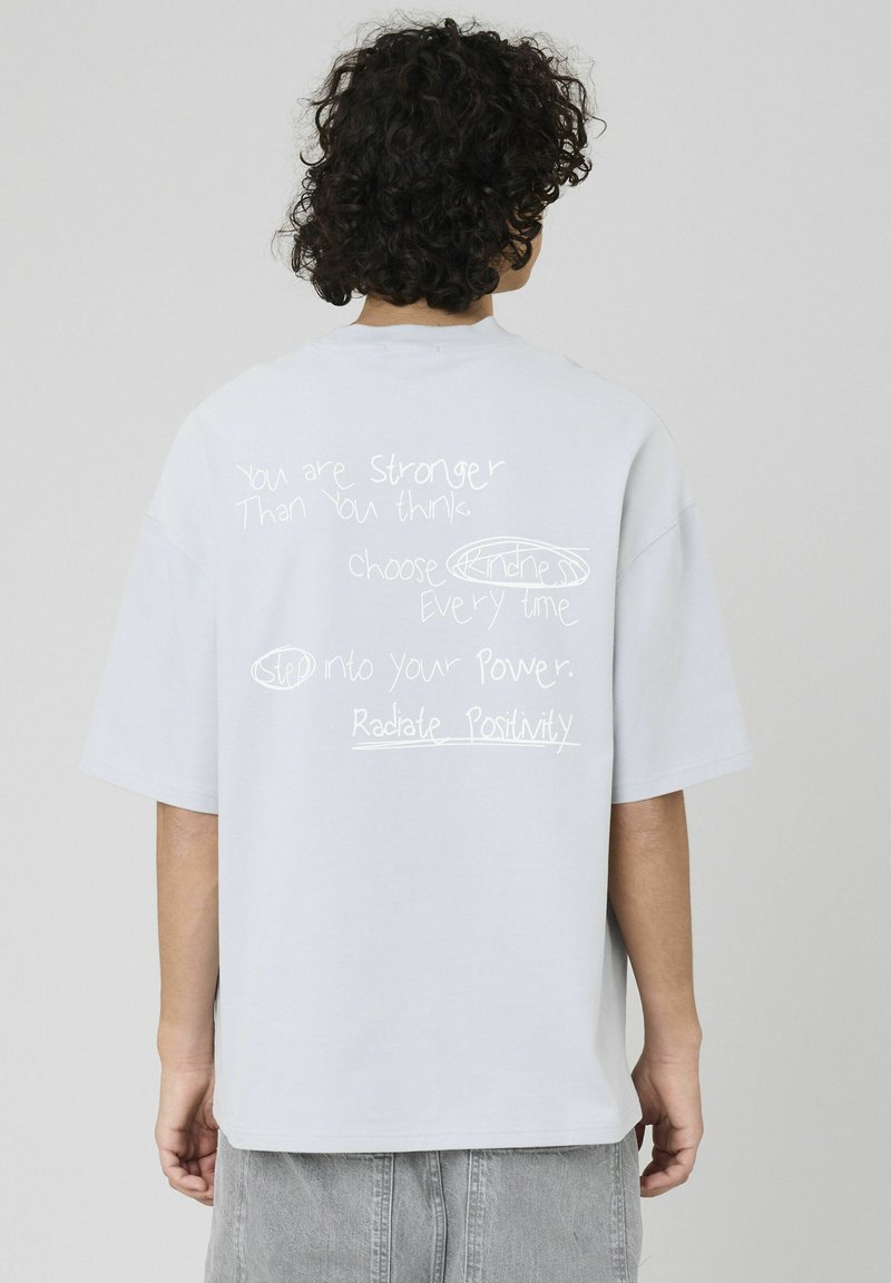 T-shirt surdimensionné gris clair à manches courtes, comportant un texte motivant manuscrit blanc au dos. Texture de tissu douce.