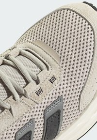 Light beige sneaker featuring a mesh upper, textured accents, and black details. Includes flat laces and a streamlined design.