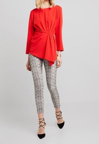 Rød asymmetrisk bluse med lange ærmer, parret med sølvfarvede slangeskinds leggings og sorte spidse flade sko med snørrebånd ved anklerne.