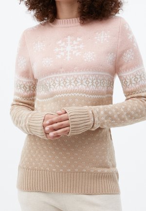 Frau trägt beige und rosa gestrickten Pullover mit weißen Schneeflockenmuster, Hände in der Taille verschränkt.