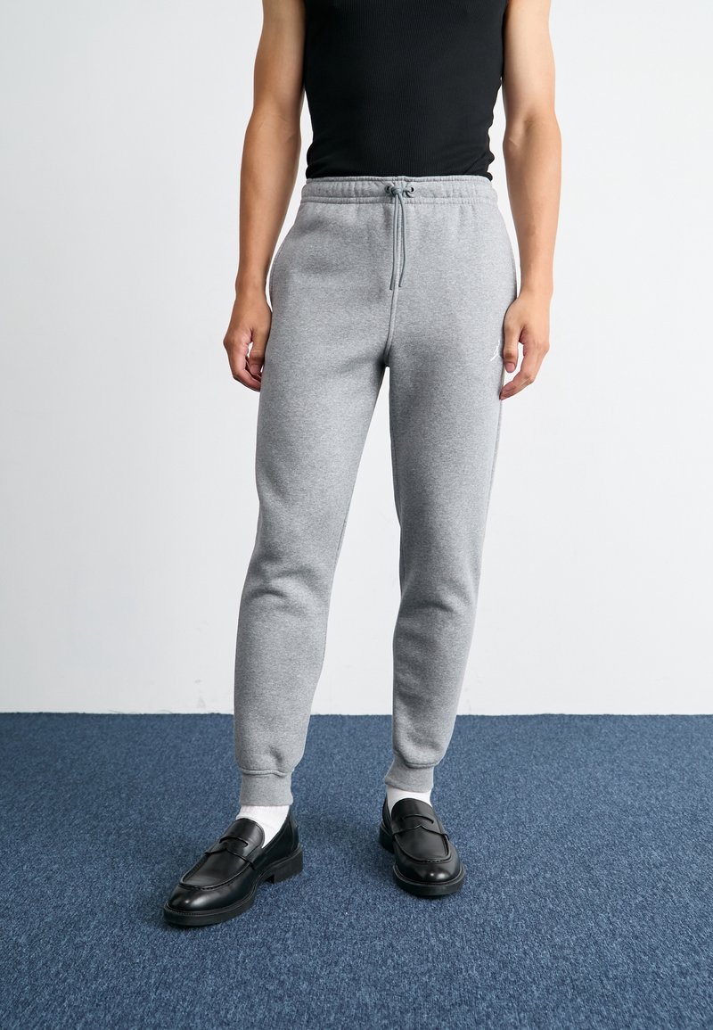 Šedé tepláky s elastickými manžetami a stahovací šňůrkou v pase, v kombinaci s černými mokasíny a bílými ponožkami. Model stojí na modré kobercové podložce.