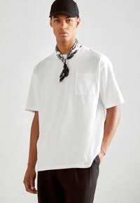 Vit kortärmad t-shirt med en bröstficka, kombinerad med svarta byxor och en svart keps. Modellen bär en mönstrad svart- och vit scarf.