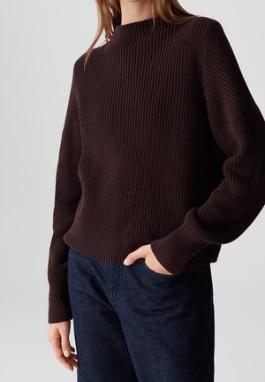 Femme portant un pull en tricot côtelé brun foncé et un jean bleu foncé, main dans la poche, sur fond clair uni.