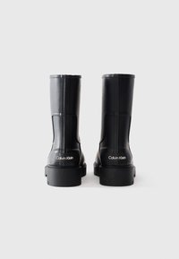 Une paire de bottes en caoutchouc noires de Calvin Klein montrée de derrière sur un fond blanc.