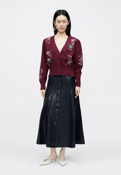 Bordó kötött kardigán virágos hímzéssel, fekete bőr gombos szoknyával párosítva. A modell törtfehér hegyes orrú cipőt visel.
