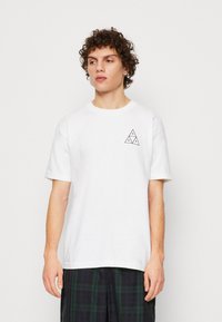 Camiseta de algodón blanca de manga corta, con un pequeño logo triangular negro en el pecho izquierdo. El modelo lleva pantalones cortos de cuadros.